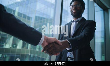 Lächelnder afroamerikanischer Geschäftsmann, der in einem Büro einem unerkennbaren kaukasischen Partner die Hand schüttelt. Ethnischer männlicher Berater, der einem Unbekannten die Hand schüttelt Stockfoto