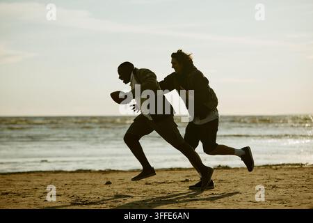 Multikulturelle Männer spielen American Football auf sandigem Strand Stockfoto