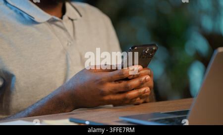 Unbekannter afroamerikanischer ethnischer Geschäftsmann, männliche Hände, die ein Mobiltelefon halten, süchtiger Benutzer, der das Gerät nachts online durchscrollt, nicht identifizierbar Stockfoto