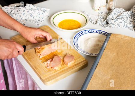 Eine Frau schneidet Hühnchen auf einem Holzbrett mit einem Messer. In der Nähe steht eine Schüssel mit geschlagenen Eiern und ein Teller mit Mehl, alles bereit für die Zubereitung von frittierten HühnerNuggets Stockfoto