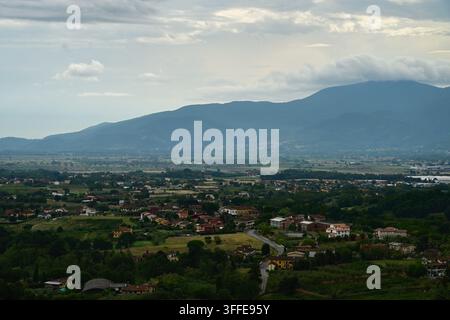 Panoramablick auf eine Stadt mit roten Dächern, Felder, eine gewundene Straße, neblige Berge und Wolken Stockfoto