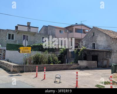 Pirovac, Kroatien, 16. August 2026 Sonnige Dorfstraße mit alten Stein- und Putzhäusern, gelbe Richtungsschilder nach Zadar und Šibenik, ruhige Straße Stockfoto