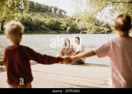 Zwei Kinder halten sich spielerisch an den Händen, während ihre Eltern im Hintergrund lächeln und an einem warmen Tag eine friedliche Uferlandschaft genießen. Stockfoto