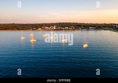 Luftaufnahme von Yachten, die bei Sonnenuntergang vor der ruhigen Küste der Insel Silba, Kroatien, vor Anker liegen. Goldenes Sonnenlicht wärmt das ruhige, tiefblaue Adriatische Meer Stockfoto