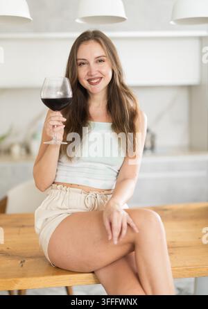 Erwachsene Frau hält ein Glas Rotwein und trinkt, während sie in der Küche steht Stockfoto
