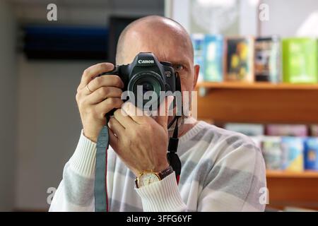 OGRE, LETTLAND - 17. FEBRUAR 2025: Männlicher Fotograf nimmt ein Spiegel-Selfie mit einer Canon-DSLR-Digitalkamera auf, die sein Gesicht im Innenraum abdeckt. Stockfoto
