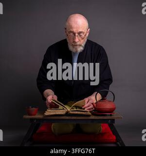 Porträt eines älteren Mannes, der einen kurzen Kimono trägt und in einer traditionellen japanischen Seiza-Position auf einem Kissen sitzt, nachdenkend, medand altes lesend Stockfoto