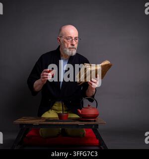 Porträt eines älteren Mannes, der einen kurzen Kimono trägt und in einer traditionellen japanischen Seiza-Position auf einem Kissen sitzt, nachdenkend, medand altes lesend Stockfoto