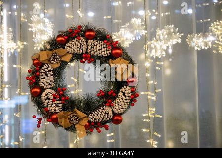 Festlicher Weihnachtskranz, der mit Tannenzapfen, roten Beeren und Ornamenten geschmückt ist, hängt vor warm leuchtenden Weihnachtslichtern mit Kopierraum. Stockfoto