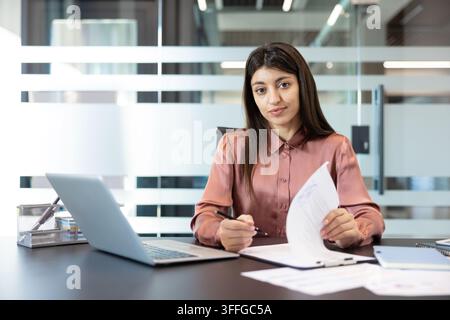 Geschäftsfrau, die Finanzberichte überprüft und Verträge auf einem Schreibtisch unterzeichnet, einen Laptop verwendet und sich auf ihre beruflichen Verantwortlichkeiten in einem Unternehmen konzentriert Stockfoto