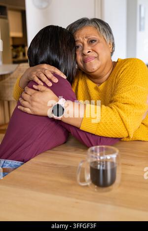 Senior Afrikanisch-Amerikanerin umarmt Familie am Küchentisch in gelbem Pullover und hält einen Glasbecher Stockfoto