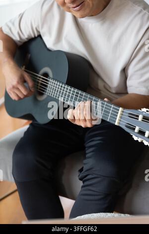 Seniorer Chinese spielt schwarze klassische Gitarre auf gepolstertem Stuhl zu Hause, Nylon Saiten Stockfoto