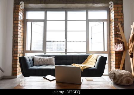 Dunkelgraues getuftetes Sofa nimmt den Mittelplatz im Loft ein, vor einem großen Gitterfenster Stockfoto
