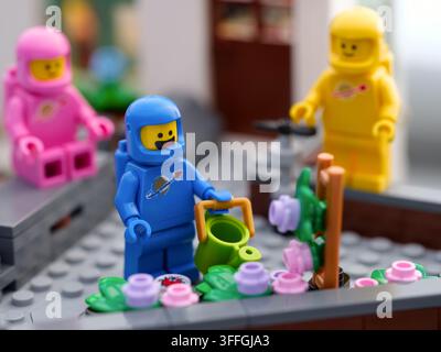 Tambow, Russische Föderation - 03. September 2026 Eine blaue Lego-Astronauten-Minifigur gießt Blumen mit einem grünen Gießkanne, während rosa und gelbe Cos Stockfoto