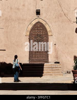 KAIRO, ÄGYPTEN: Eine Frau mit einem Rucksack geht an der monumentalen, mit Kreuz geschnitzten Holztür einer historischen koptischen Kirche im alten Kairo vorbei. Vertikal. Stockfoto