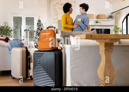 Zwei afroamerikanische Männer umarmen sich und lächeln im Wohnzimmer mit Koffern und Weihnachtsbaum Stockfoto