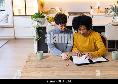 Zwei afroamerikanische Männer sitzen an einem Holzküchentisch und unterschreiben Papiere in einem schwarzen Portfolio mit einem Stift Stockfoto