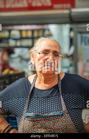 Seniorische Straßenhändlerin mit Brille und Schürze. Porträt einer älteren Marktfrau, die an einem Außengastronomiestand in die Kamera schaut. Stockfoto