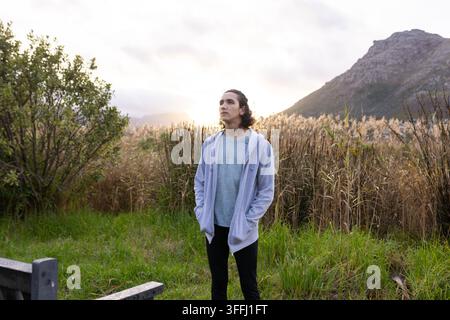 Mann trägt einen hellgrauen Kapuzenpullover mit Reißverschluss, ein hellblaues T-Shirt, steht auf einer Wiese in der Nähe einer Bank, Schilf, Gegenlicht Stockfoto