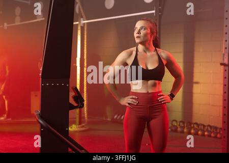 Frau Mitte 20 in einem dämmrigen Fitnessstudio, trägt einen schwarzen Sport-BH und rote Leggings mit hoher Taille Stockfoto