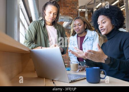 Afroamerikanische Kollegen, die an einem Holztisch neben dem Fenster auf einem Laptop zeigen, einer mit Rollkragenpullover Stockfoto