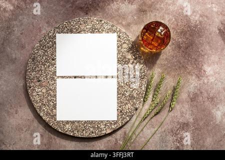 Whisky-Glas mit Eis, leere weiße Visitenkarten-Vorlage mit Elementen aus Granit und Weizenähren. Männlicher Hintergrund. Alkoholindustrie-Vorlage. Oberansicht, Flat Lay. Beige Grunge-Hintergrund Stockfoto