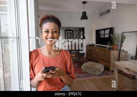 Lächelnde Frau, die ein Smartphone benutzt und am Fenster zu Hause steht Stockfoto