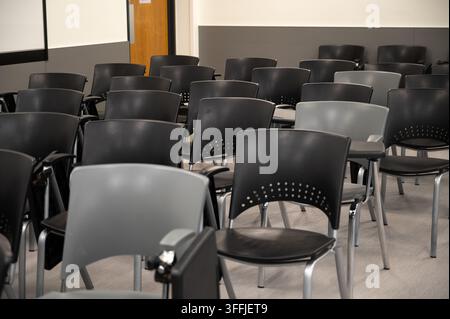 Reihen von leeren schwarzen und grauen Stapelsesseln mit Tablet-Armlehnen stehen einem an der Wand montierten Projektionsbildschirm und einer Holztür in einem Hörsaal auf der g Stockfoto