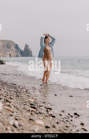 Strandfrau im Urlaub, die auf einer Kieseluferpromenade mit Meereswellen und malerischen Felsformationen unter einem weichen Himmel spaziert Stockfoto