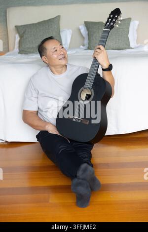 Älterer chinesischer Mann sitzt auf Holzboden am Fuß des Bettes, hält eine schwarze Gitarre und trägt eine Armbanduhr Stockfoto