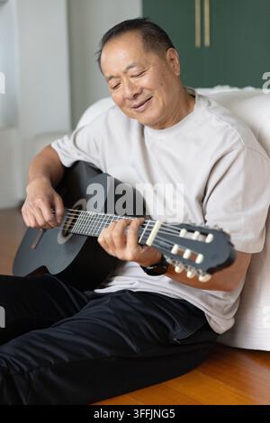 Seniorer asiatischer Mann sitzt auf Hartholzboden und spielt schwarze Akustikgitarre neben einem weißen Sofa zu Hause Stockfoto
