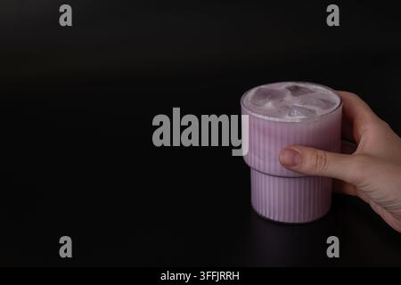 Hand, die ein klares Glas mit pinkem Matcha und Eiswürfeln gegen einen dunklen Hintergrund hält, wodurch eine erfrischende Nahaufnahme einer Getränkeszene mit natürlichem Licht entsteht Stockfoto