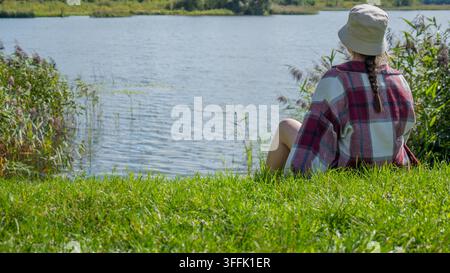 Frau mit einem leichten Sonnenhut und kariertem Hemd sitzt an einem grasbewachsenen Seeufer mit Blick auf das ruhige Wasser. Der offene Platz neben ihr schafft Platz für Text, während der Stockfoto