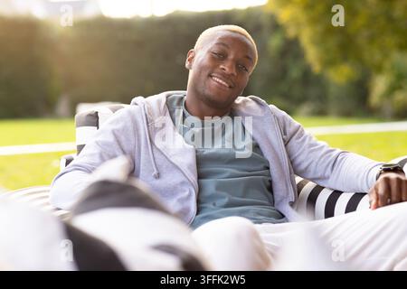Afroamerikaner sitzt zurückgelehnt auf einem gestreiften Stuhl auf einer Rasenfläche und trägt einen Hoodie Stockfoto