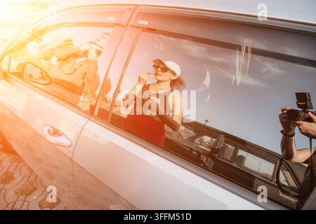 Fensterreflexion eines Autos einer jungen Frau, die Lippenstift aufträgt, während sie draußen auf ihr Telefon schaut Stockfoto