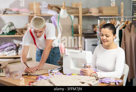 Team von Schneidern, die in der Werkstatt arbeiten. Asiatische Schneiderin näht Kleidung neben einem Herrenmodedesigner, der Muster auf Papier zeichnet Stockfoto