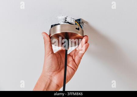 Ein Mann positioniert den runden, gebürsteten Metallschirm einer Deckenleuchte über ihrer Montagehalterung, wobei oben noch freiliegende Verkabelung sichtbar ist und der schwarze Fab Stockfoto