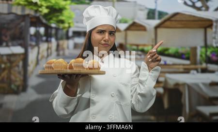 Die junge Küchenchefin hält ein Tablett mit Muffins und zeigt mit dem Finger an den Verkaufsständen auf dem Straßenmarkt; Selbstvertrauen stolz auf Handwerkskunst Wärme. Stockfoto