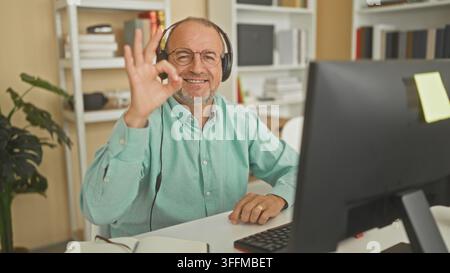 Mann mit Headset macht Ok-Handgestik vor Computermonitor im Bürogebäude, Callcenter-Agent sitzt; freundliche Unterstützung. Stockfoto
