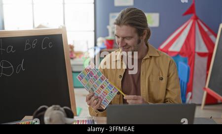 Mann Lehrer zeigt Bleistift auf bunte Alphabetkarte neben Tafel im Vorschulgebäude; frühe Alphabetisierung Wärme. Stockfoto