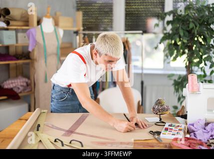 Junger männlicher Schneider zeichnet ein Muster auf ein Blatt Papier mit Kurven, während er ein Kleid an seinem Arbeitsplatz in einer Mode-Nähwerkstatt näht Stockfoto