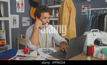 Mann mit Messband, der das Kinn auf die Hand stützt, während er einen Laptop benutzt und Skizzen im Studio begutachtet; nachdenkliche kreative Konzentration. Stockfoto