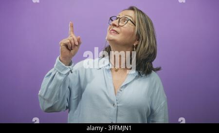 Frau hispanic Senior mit grauem Haar und Brille zeigt den Zeigefinger nach oben, während sie vor einem violetten Studio-Hintergrund aufblickt; Neugier. Stockfoto