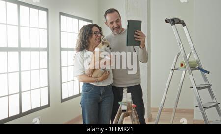 Mann und Frau, mittelaltes Paar, lächeln, während die Frau einen kleinen Hund hält und die Hand des Mannes ein Tablet hält, um ein Selfie in einem Gebäude zu machen; Freude an der Hausrenovierung. Stockfoto