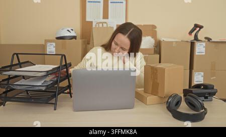 Frau am Schreibtisch sortiert Pakete und Papierkram neben einem Laptop, greift nach einem Dokumentenbehälter und hält die Hand an den Hals in einem Verpackungsgebäude; E-Commerce-Erschöpfung Stockfoto