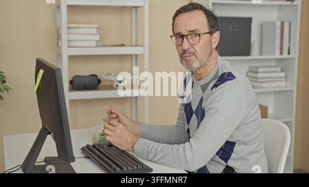 Mann hält Gabel zum Mund, isst Salat am Computer in einem Gebäude, Hände auf Schüssel und Tastatur, trägt Brille und Argyle-Pullover, während er am Schreibtisch sitzt; con Stockfoto