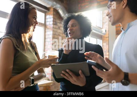 Verschiedene Kollegen diskutieren ein Projekt in einem Loft-Büro, halten ein Tablet, ein Smartphone und einen Glastopf Stockfoto