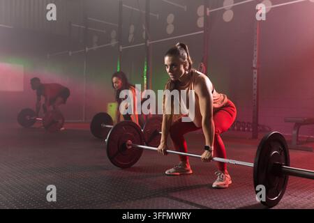 Verschiedene Klassenkameraden in Sportkleidung heben belastete Hantelstangen in einem dämmrigen Fitnessstudio mit Kettlebells Stockfoto