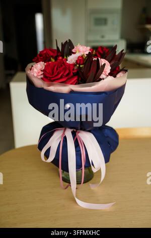 Strauß mit roten Rosen und pinken Nelken, in marineblauem Papier eingewickelt und mit hellrosa Bändern gebunden, auf einem Tisch stehend, romantisches Geschenk, Blumenarrangement, Jubiläumsfeier Stockfoto