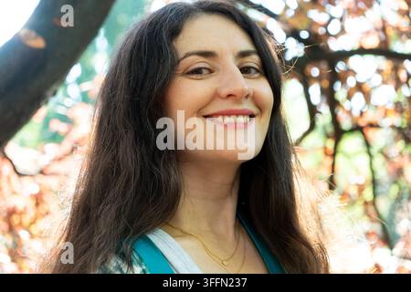 Gelassene Frau mit langen lockigen Haaren blickt im sonnigen Naturpark in die Kamera Stockfoto